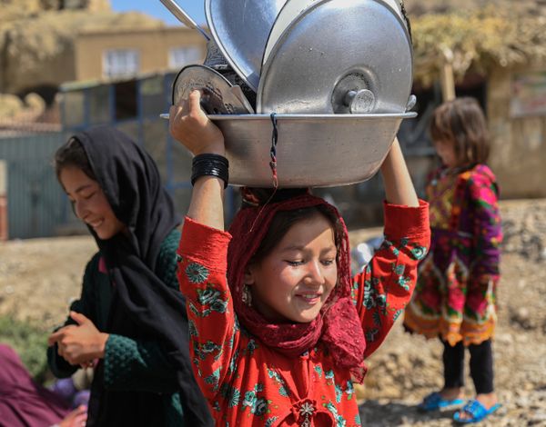 Eine junge afghanische Frau trägt Kessel auf dem Kopf und lächelt konzentrirt, dahinter steht eine zweite junge Frau.