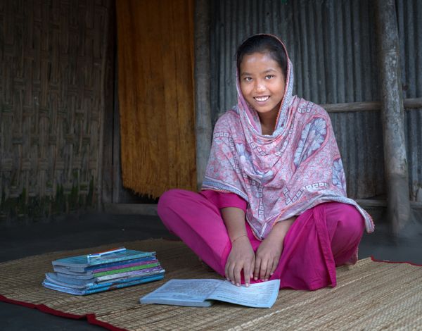 Eine junge Frau sitzt am Boden, vor sich ein Buch, und lächelt in die Kamera.