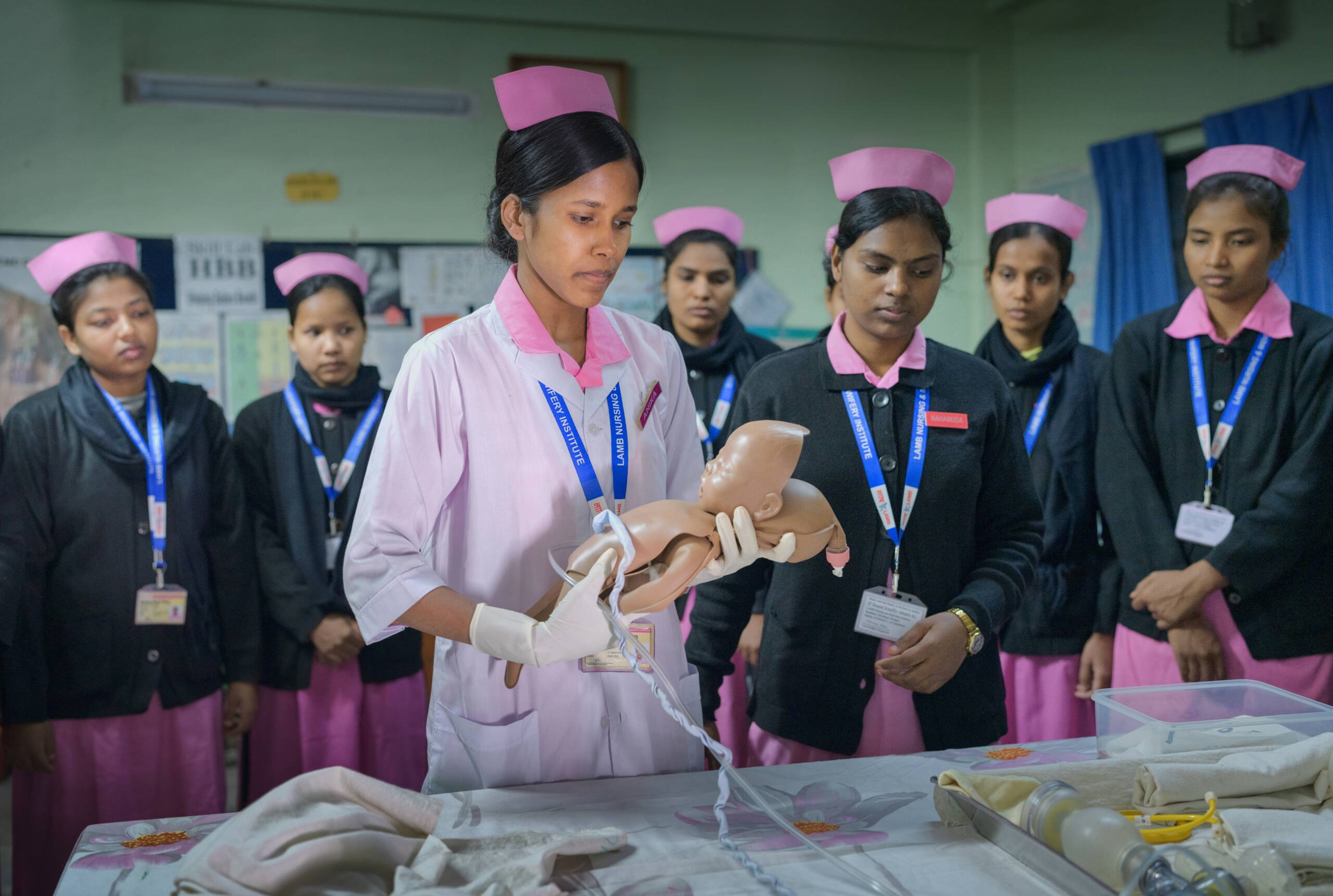 Hebammenausbildung Junge Frauen stehen an einem Tisch, eine hält eine Babypuppe hoch.