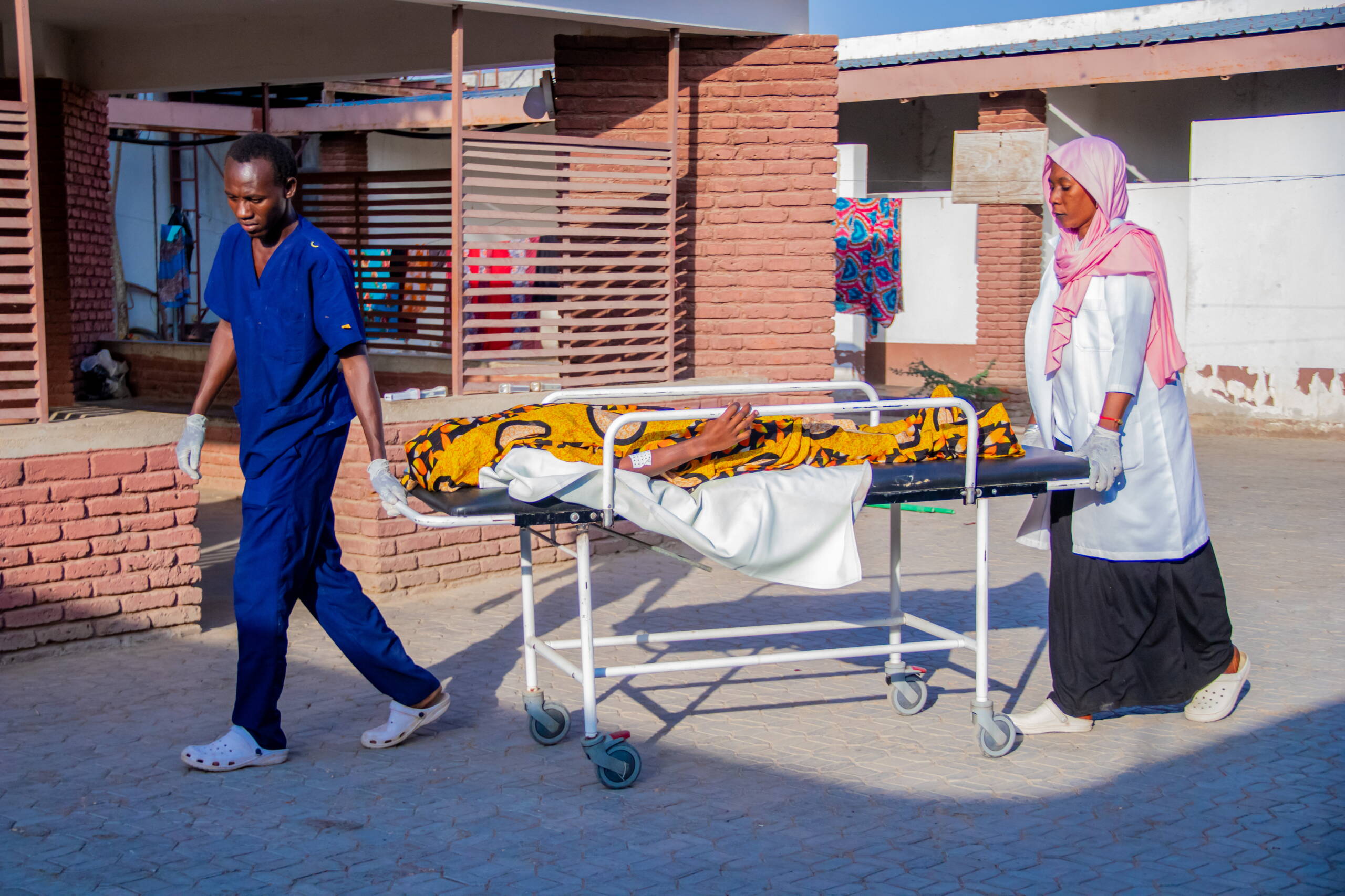 Zwei medizinische Fachpersonen schieben eine Frau auf einer Liege über einen Innenhof.