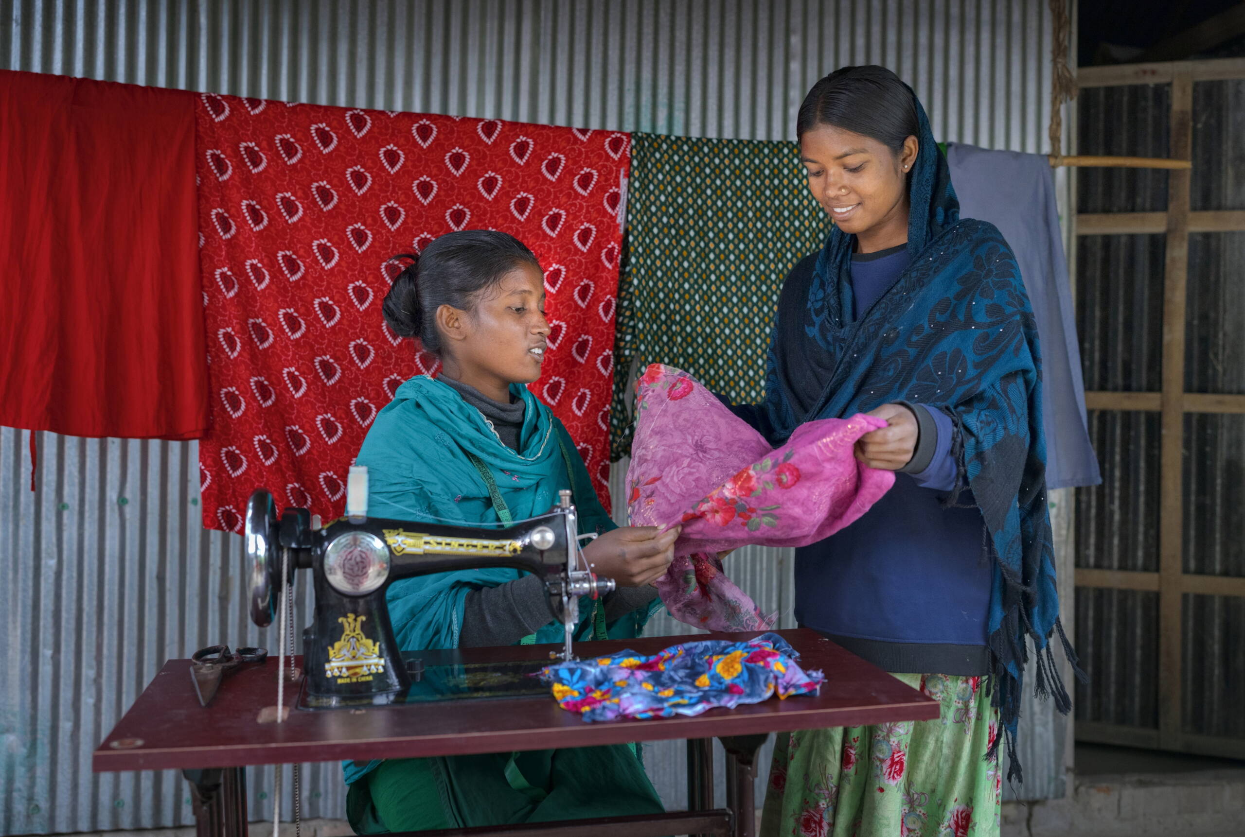 Ein Mädchen sitzt an einer Nähmaschine, ein anderes steht daneben. Gemeinsam betrachten sie ein Stück Stoff.