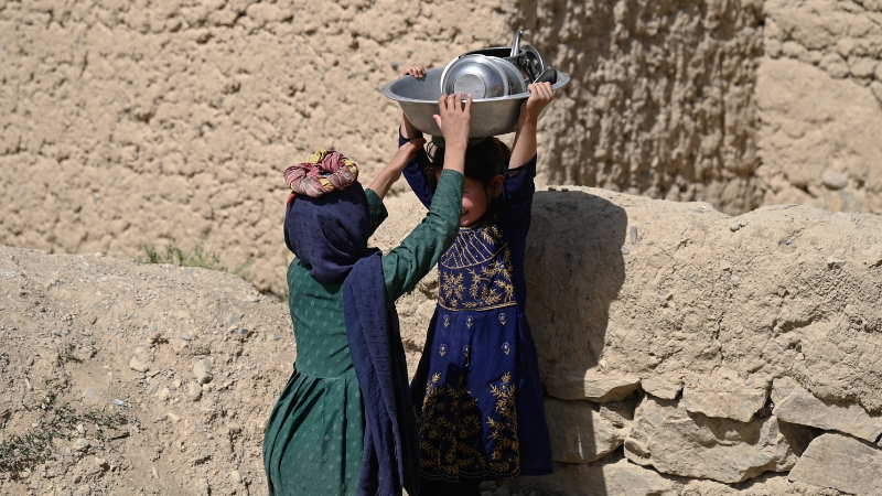 Mädchen Afghanistan Zwei afghanische Mädchen stehen einander gegenüber, eine hält Geschirr auf dem Kopf, das andere greift danach.