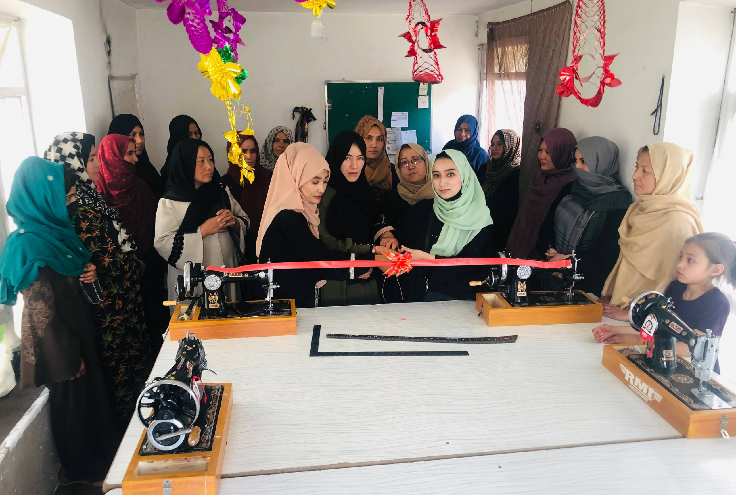 Zwei Frauen schneiden ein Band durch, das zwei Nähmaschinen verbindet, dahinter stehen verschiedene Frauen, von der Decke hangen Girlanden.