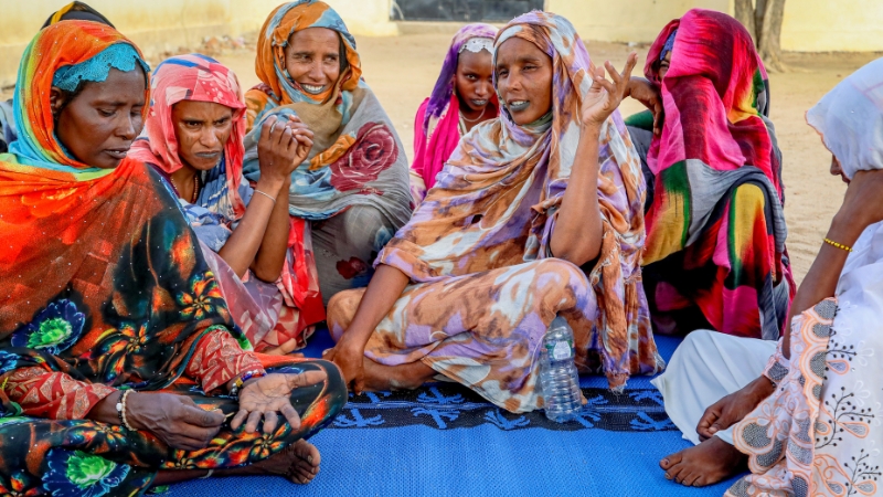 Frauen in farbigen Kleidern sitzen am Boden. Eine hebt die Hand, um etwas zu sagen.