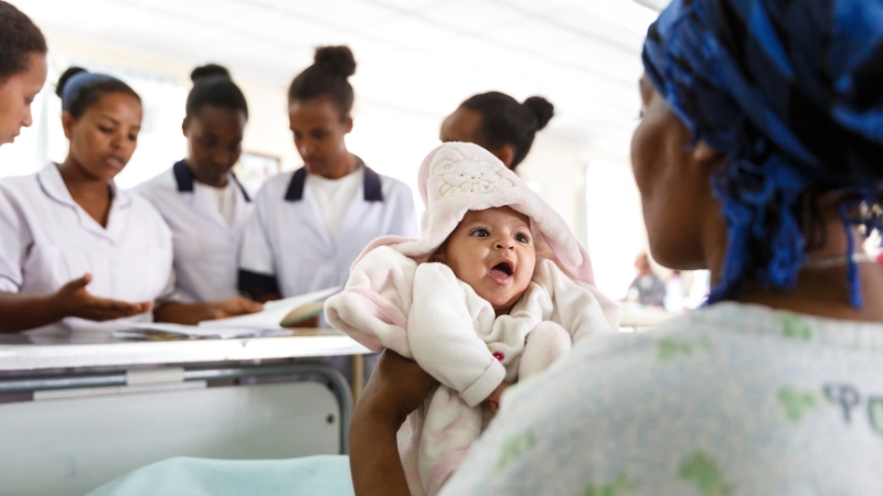 Eine Hebamme für jede Mutter Eine Hebamme hält ein Neugeborenes hoch, dahinter weitere Fachfrauen im Gespräch.