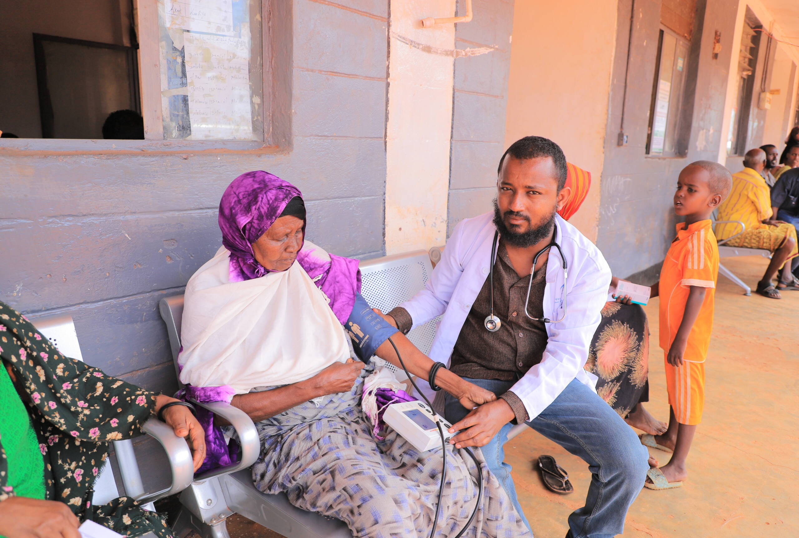 Ein Mann misst einer Frau den Bluckdruck, sie sitzen draussen vor einem Gebäude.