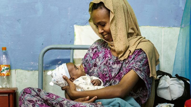 Eine Mutter sitzt auf dem Spitalbett und schaut ihr Neugeborenes an.