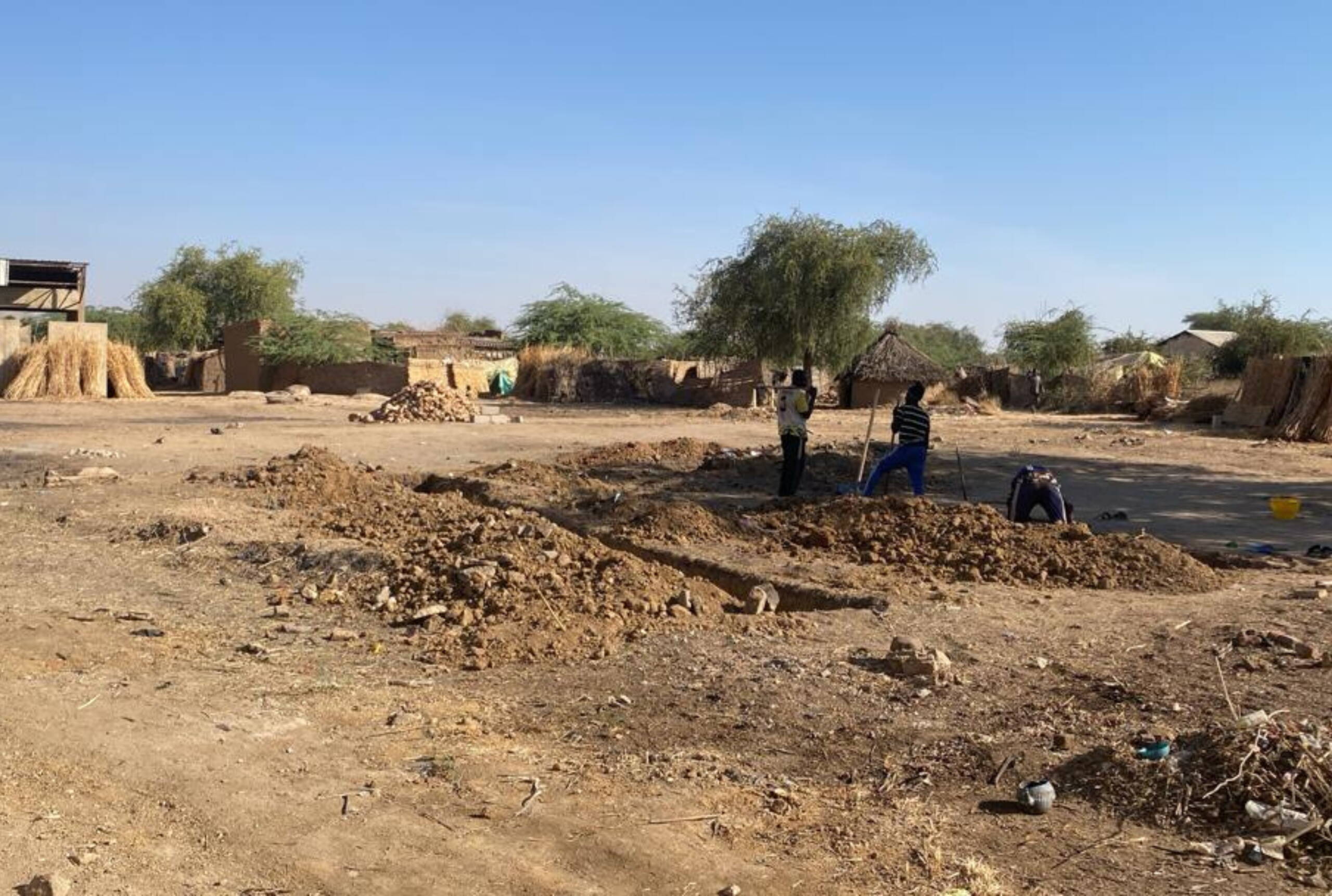 Sandige Fläche mit Häusern im Hintegrund, drei Männer beginnen, den Boden auszuheben.