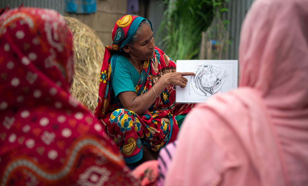 Eine Frau erklärt anderen Frauen etwas und hält eine Illustration mit einem Querschnitt von einem schwangeren Unterleib in den Händen.