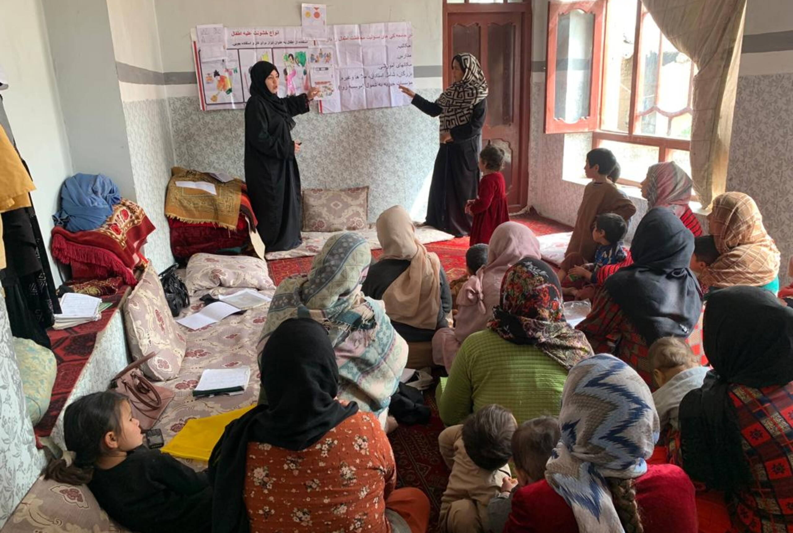 Zwei Frauen erklären etwas und zeigen auf Illustrationen an einer Wand; davor sitzen Frauen am Boden und hören zu.