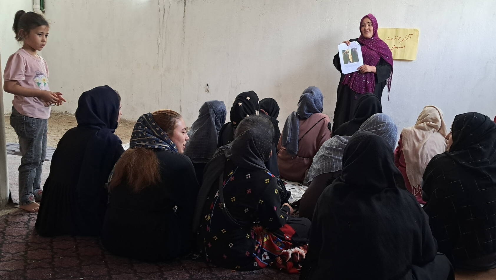 Frauen sitzen am Boden und hören einer Frau zu, die ein Bild in die Höhe hält.