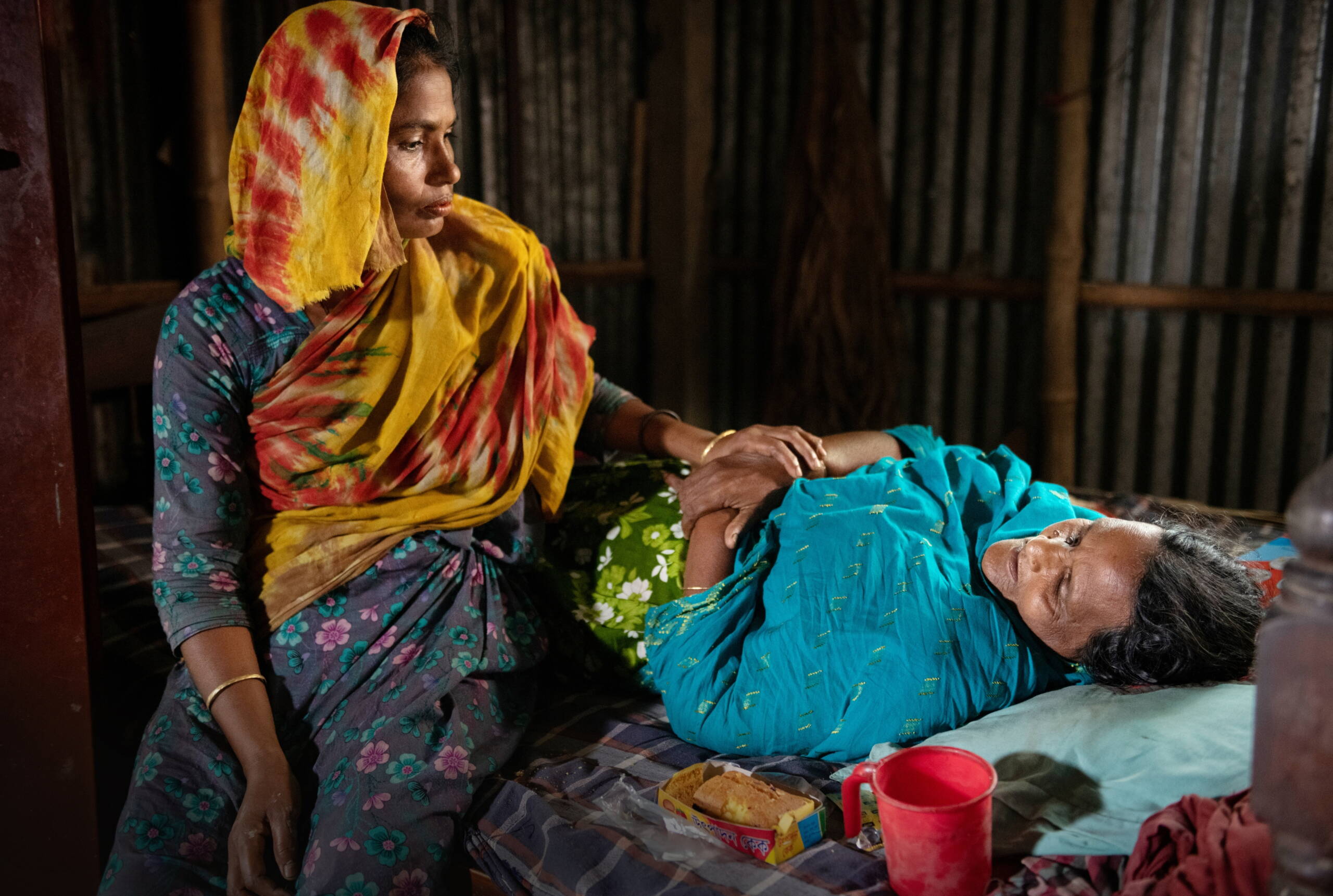 Eine Frau liegt auf einem Bett, eine zweite Frau hält deren Hand.