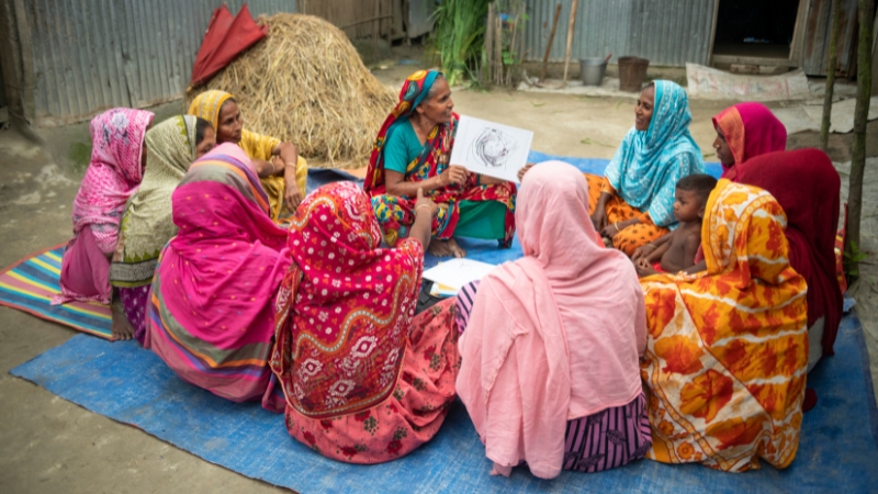 Frauen sitzen am Boden im Kreis, eine Frau erklärt etwas und hält eine Grafik in den Händen.