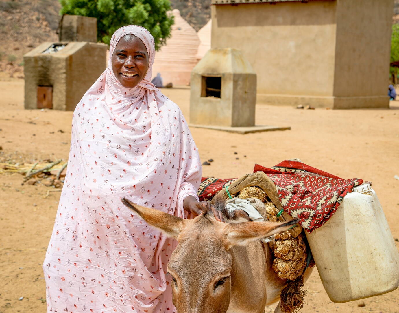 Jetzt spenden! Eine schwangere Frau mit Esel an ihrer Seite steht vor ein paar Gebäuden und lächelt in die Kamera.