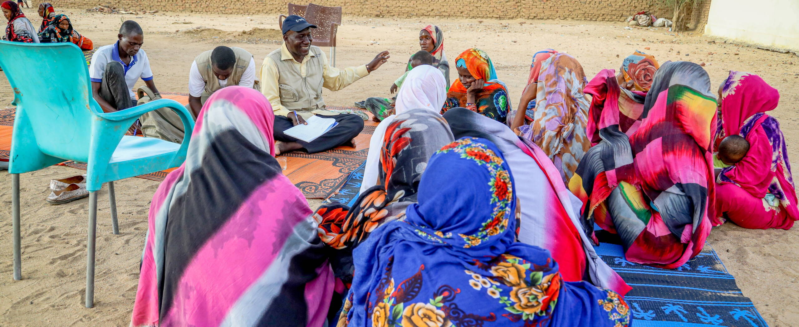 Frauen sitzen am Boden und werden von einem Mann befragt. Dieser lacht und hält die Hand hoch.