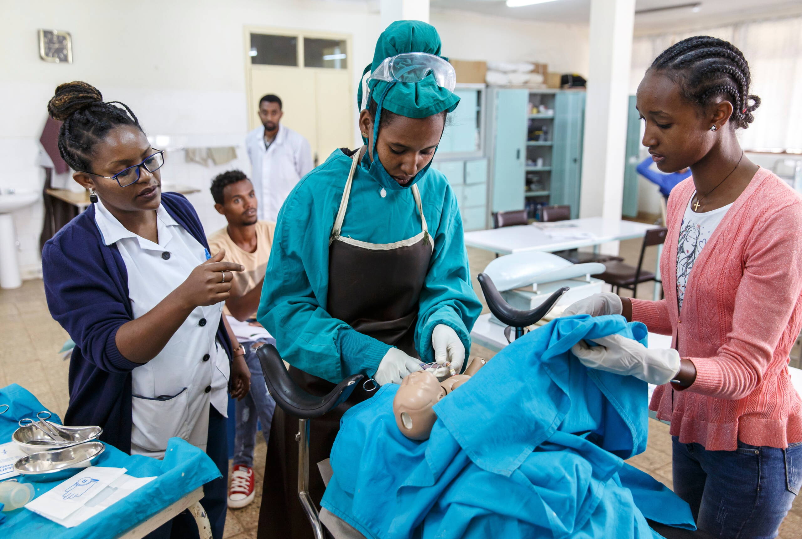Hebammenausbildung Eine Frau in Schutzkleidung arbeitet mit einer Babypuppe, zwei Frauen schauen zu.