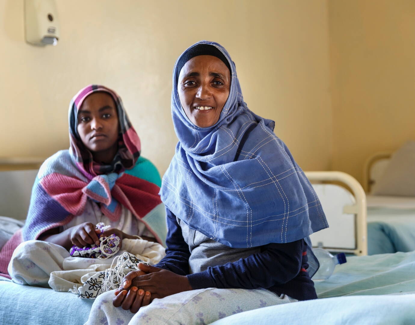 Zwei Frauen sitzen auf einem Spitalbett und schauen in die Kamera.