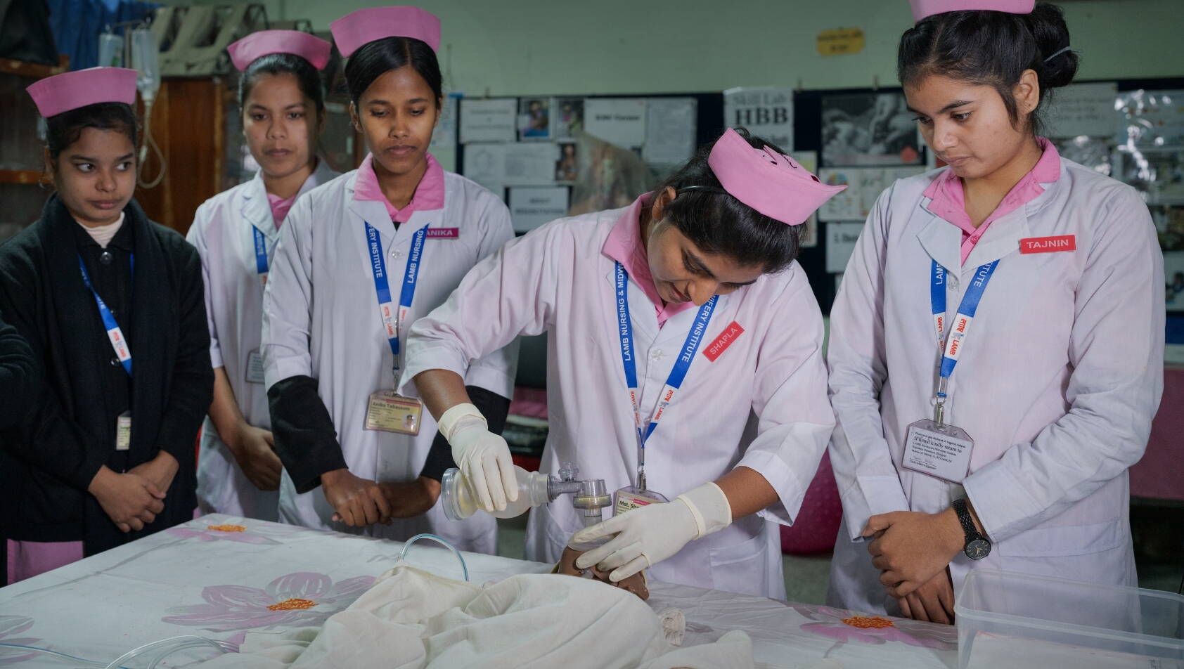 Hebammenstudentinnen stehen an einem Tisch, eine junge Frau drückt eine Beatmungsmaske auf eine Babypuppe.