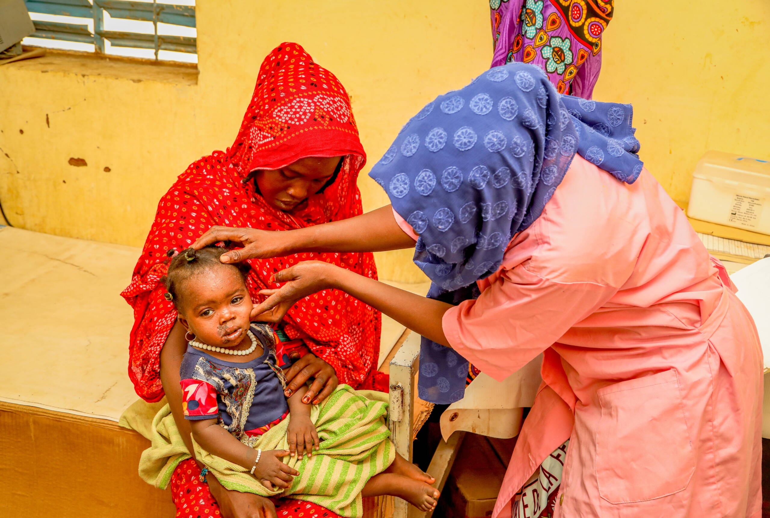 Eine Hebamme untersucht ein Kleinkind, das bei seiner Mutter auf der Schoss sitzt.